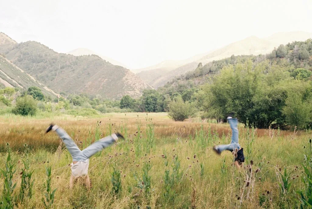 Two people doing cartwheels in a grassy field with forested mountains in the background