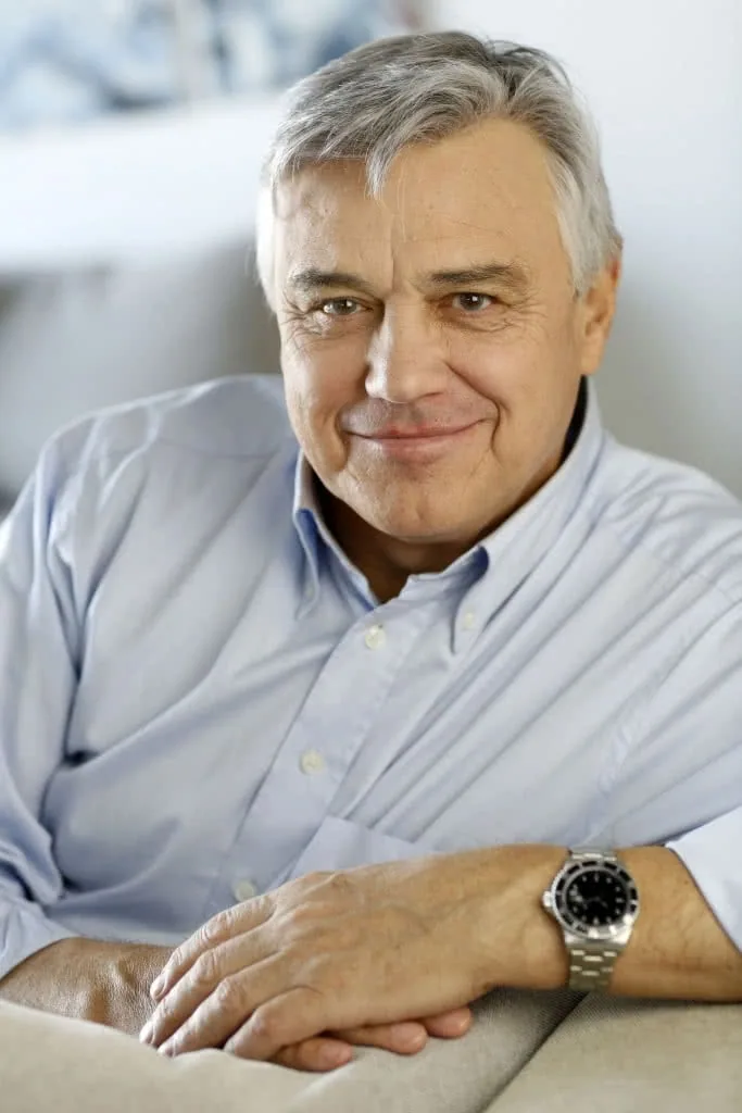 Older man with gray hair wearing light blue button-up shirt and watch, smiling at camera in professional portrait