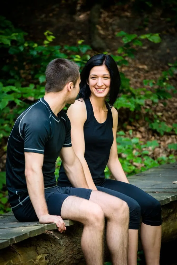Two people in athletic wear sitting together on a wooden bench outdoors with green foliage in background