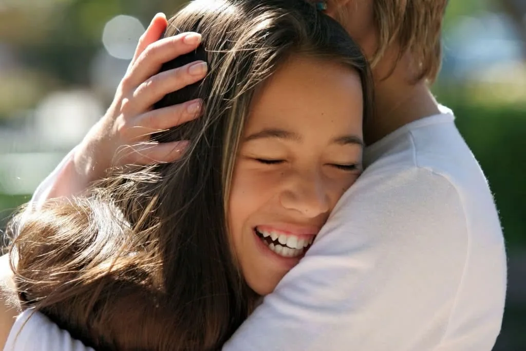 Woman smiling while being embraced from behind by a person in a white shirt outdoors