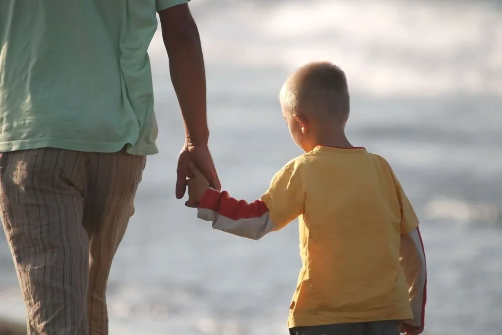 Adult and young child holding hands while standing on beach facing ocean waves