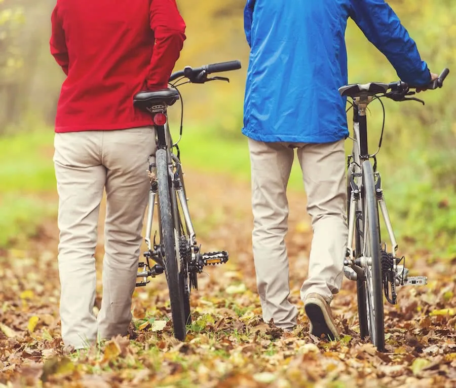 Two people wearing red and blue jackets standing with bicycles on a leaf-covered ground outdoors.