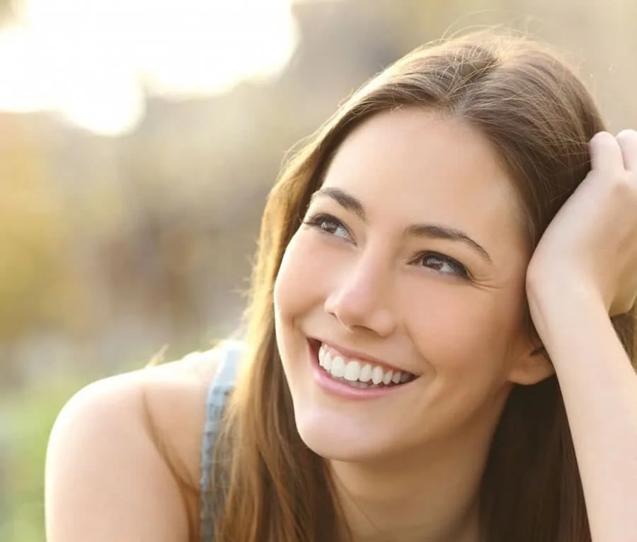 Woman smiling outdoors with hand near head and blurred natural background