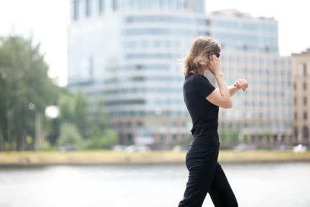 42259184 - young confident business woman walking in hurry, looking at watch, talking on mobile phone on the city street in front of blue glass modern office building beside riverbank, profile view