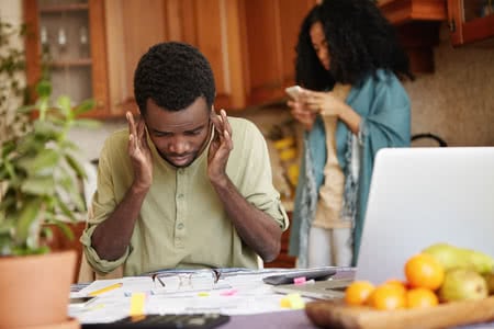 71992997_s 71992997 - young african man plugging his ears, feeling stressed, can't stand financial stress anymore, not able to cover his debts while managing family budget at kitchen table with laptop and lots of papers