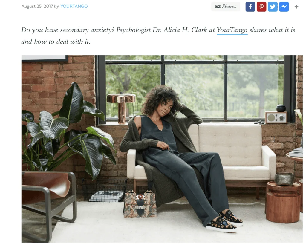 Woman with curly hair sitting on a beige couch in a brick-walled room with plants and windows