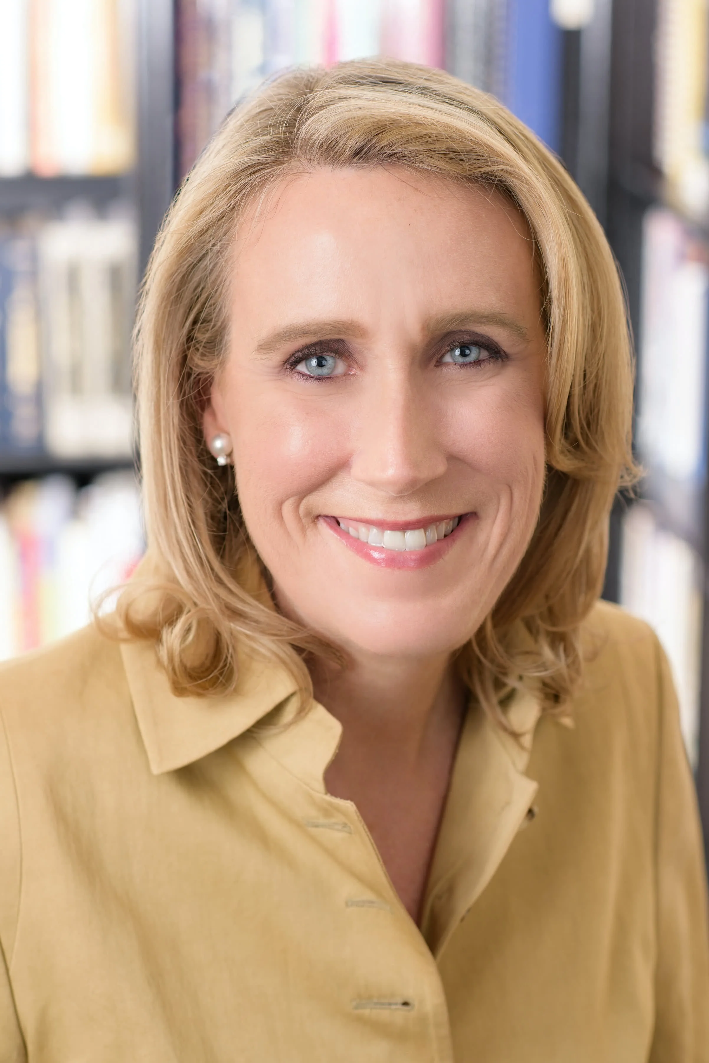Woman with shoulder-length blonde hair wearing a yellow collared shirt, smiling at camera with bookshelves blurred in background.