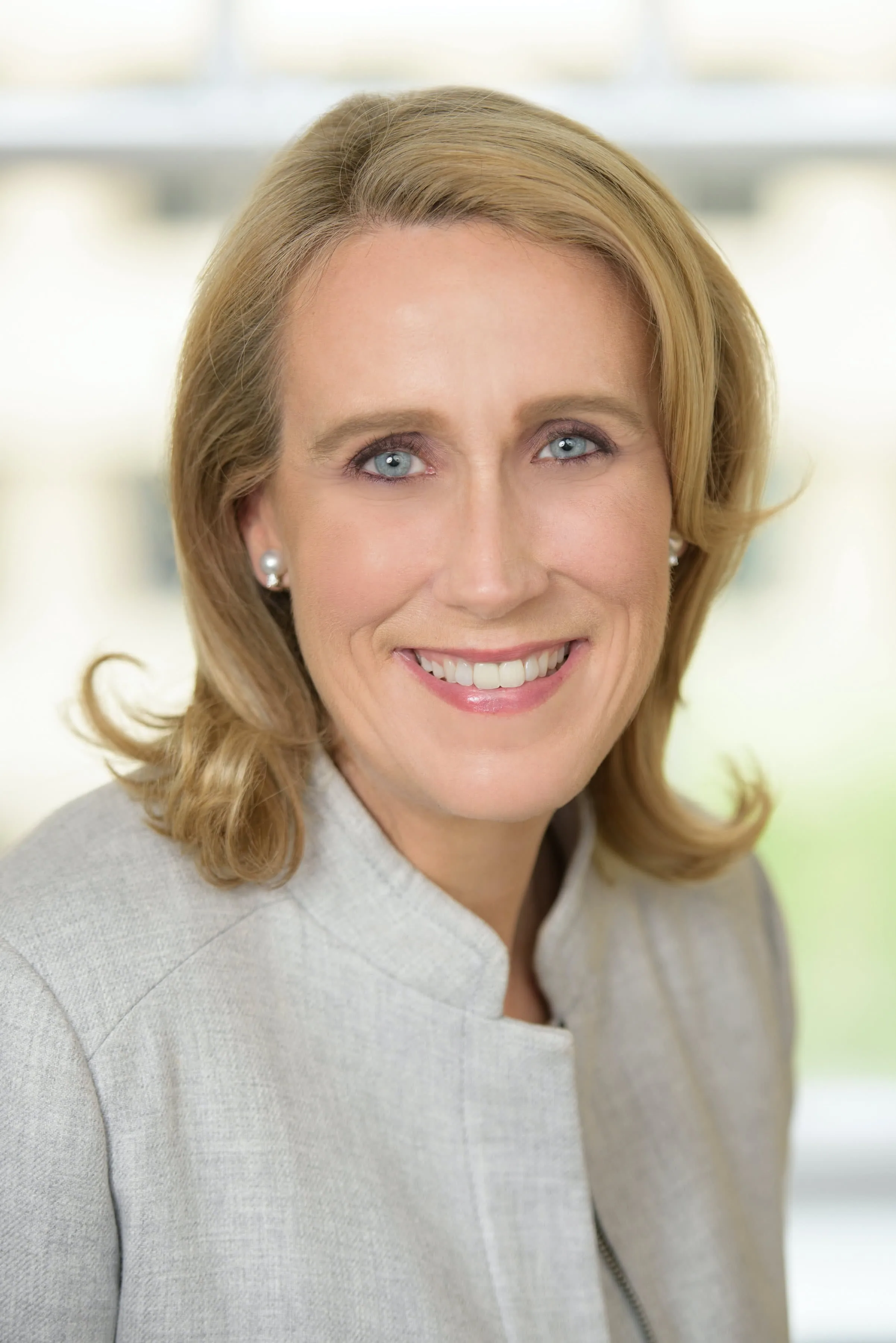 Professional headshot of a woman with shoulder-length blonde hair wearing a gray blazer and pearl earrings, smiling at the camera