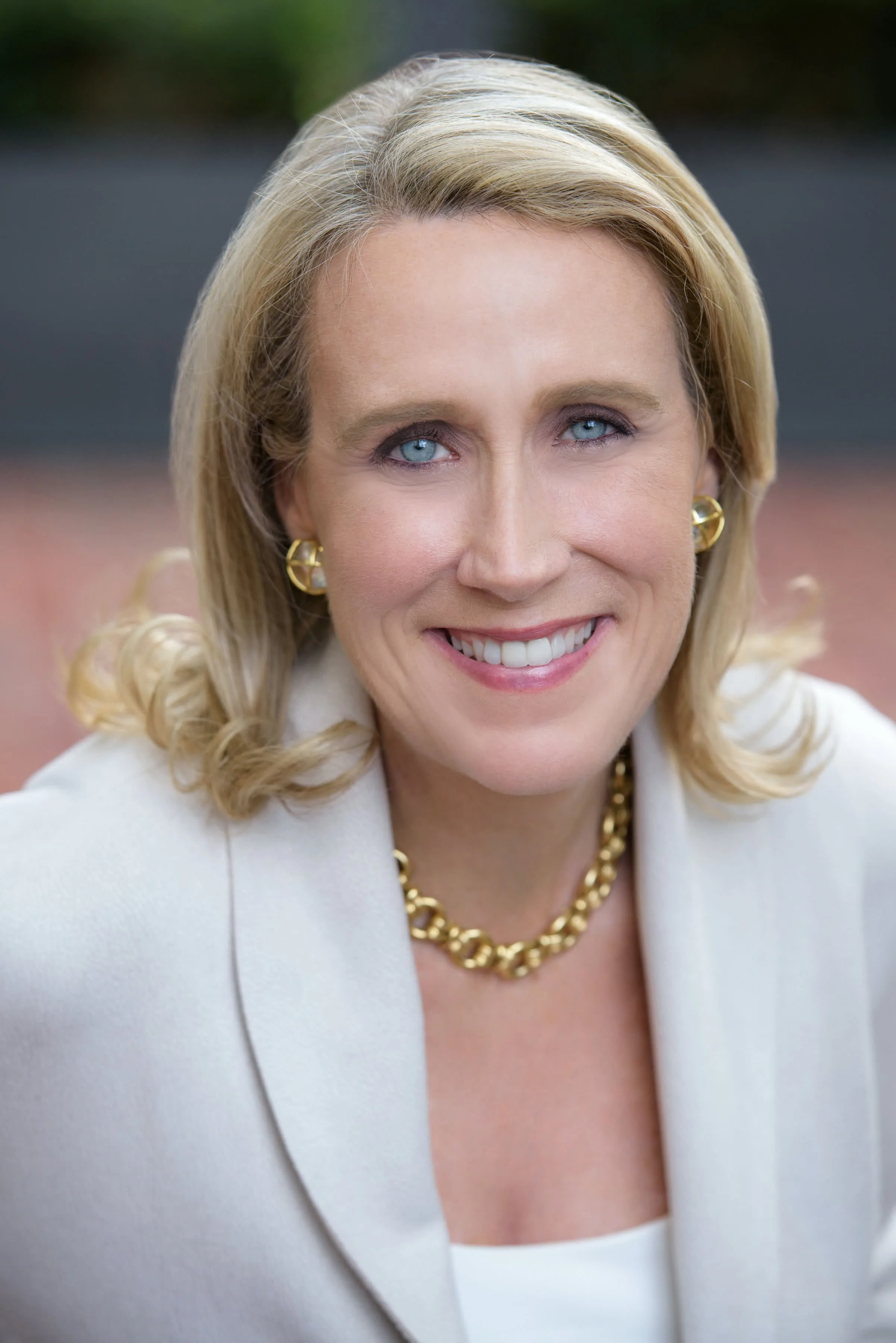 Professional headshot of a woman with shoulder-length blonde hair wearing a cream blazer, gold necklace, and gold earrings, smiling at camera