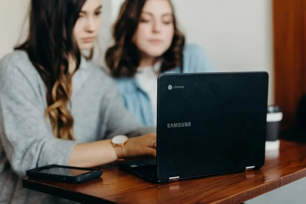 Two women viewing a black Samsung Chromebook on a wooden table with a smartphone beside it