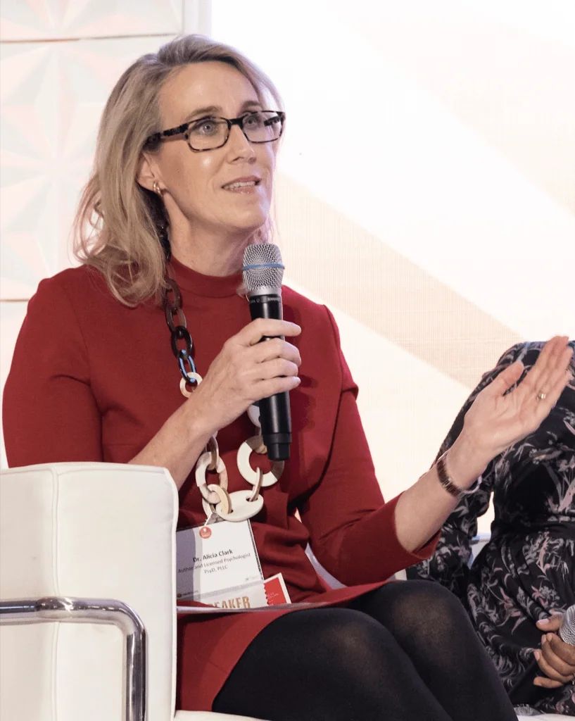 Woman wearing red top and glasses speaking at event while holding microphone