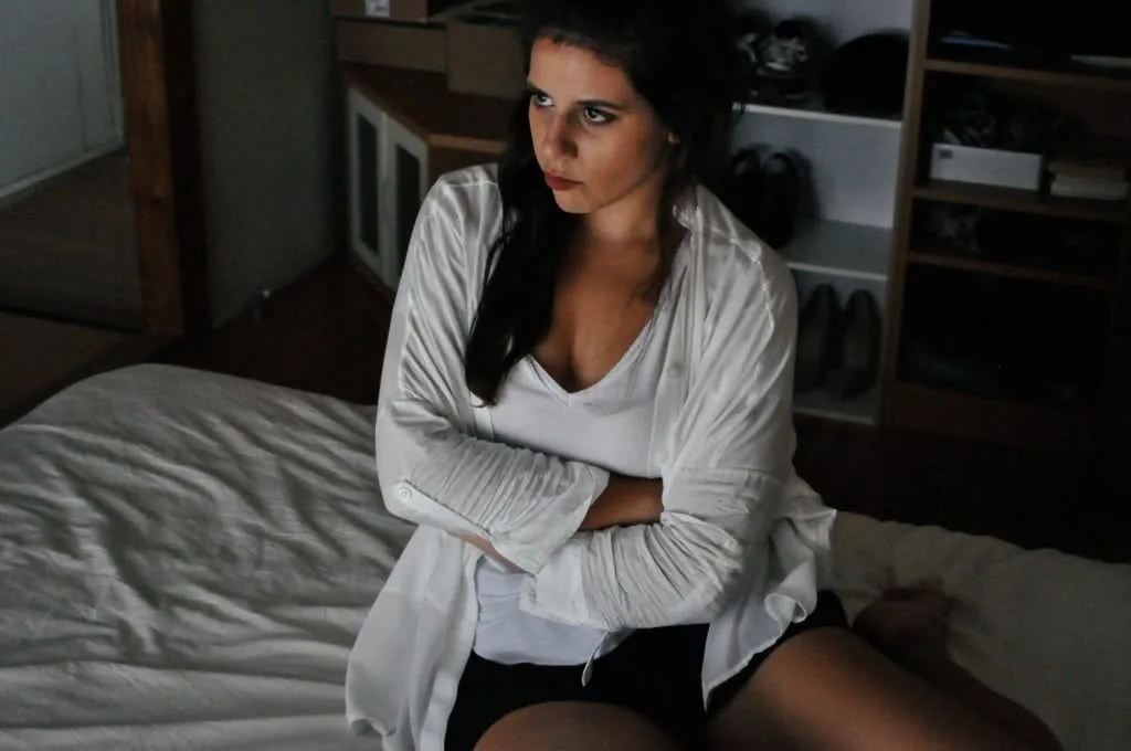 Woman sitting on bed in bedroom wearing white cardigan over white top, looking downward with troubled expression