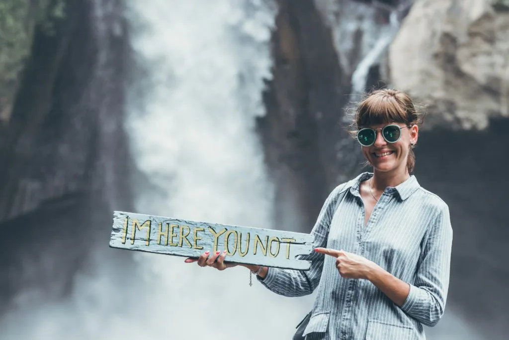 Woman wearing sunglasses holding wooden sign reading 'I'M HERE YOU NOT' in front of waterfall